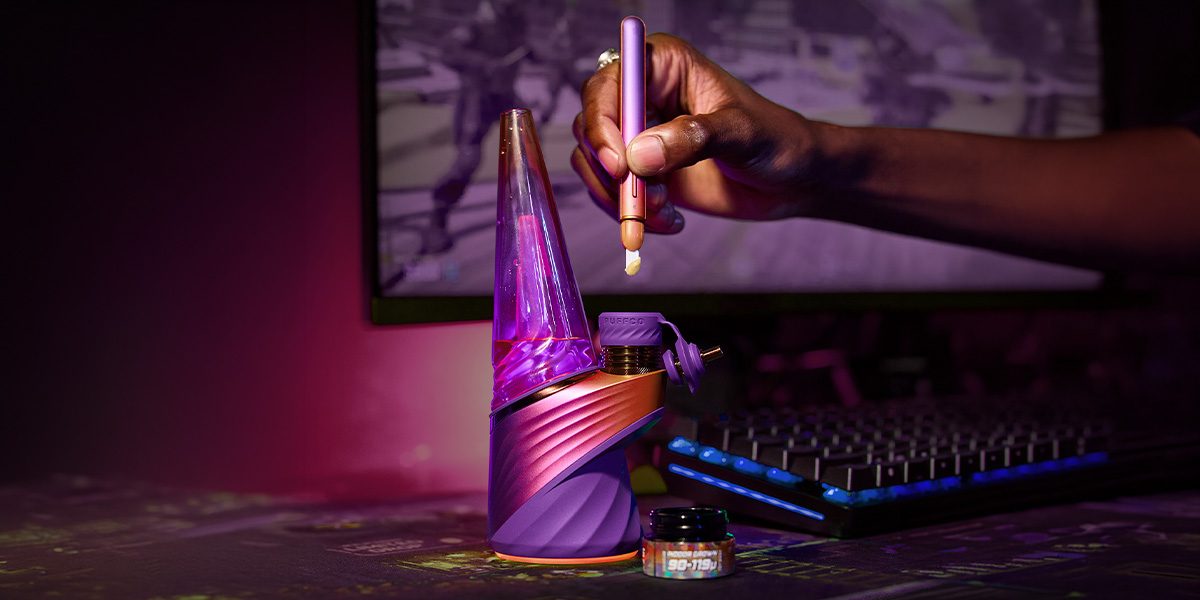 Puffco Plasma Edition Smart Rig im Gaming-Setup, während eine Hand Konzentrat mit einem Dab-Tool einfüllt.