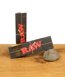 Zwei Packungen RAW Black King Size Papers auf einem Stein. Hochwertige, ungebleichte Blättchen für puren Geschmack.
