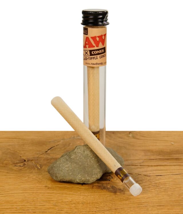 Zwei RAW DLX Glass Tipped Cones, einer im Glasröhrchen und einer daneben auf einem Stein liegend, auf einem Holzuntergrund.