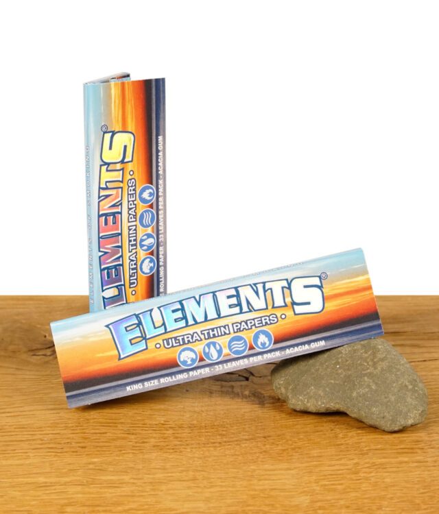 Zwei Packungen Elements King Size Ultra Thin Papers auf einem Holztisch, eine liegend und eine stehend, zeigt das farbenfrohe Design der Verpackung.