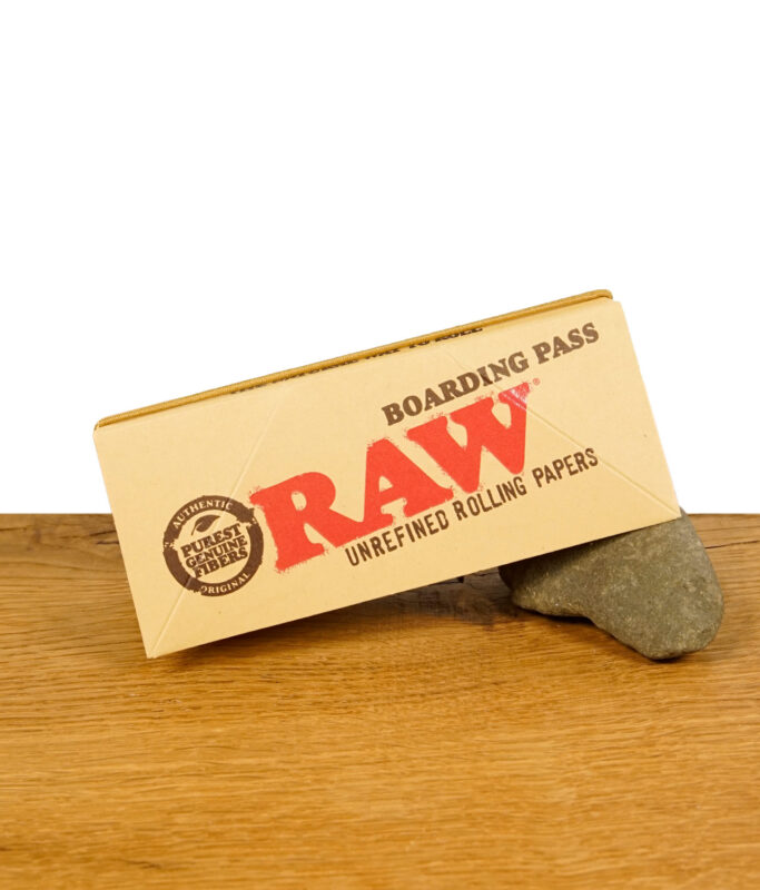 Der geschlossene RAW Boarding Pass auf einem Stein liegend, zeigt das RAW-Logo und den Text 'Unrefined Rolling Papers'.
