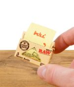 Hand hält RAW Classic Rolls Single Wide Zigarettenpapierpackung, zeigt Ziehanweisung.