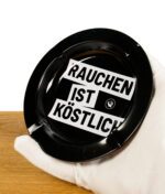 Schwarzer Aschenbecher auf Holzbrett mit Rauchen ist Köstlich Spruch bedruckt von einer Hand gehalten