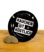 Schwarzer Aschenbecher auf Holzbrett mit Rauchen ist Köstlich Spruch bedruckt