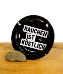 Schwarzer Aschenbecher auf Holzbrett mit Rauchen ist Köstlich Spruch bedruckt