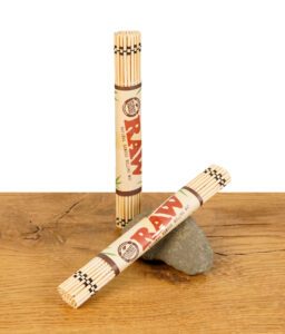 RAW Bamboo Rolling Mat, zusammengerollt und mit Papierbanderole, stehend und liegend auf Holztisch mit Stein.