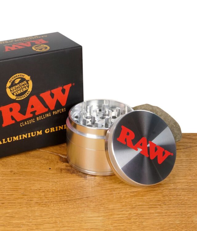 Geöffneter RAW Aluminium Grinder mit Verpackung im Hintergrund, Deckel mit rotem RAW-Logo, auf Holztisch mit Stein.
