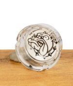 Nahaufnahme des The Bulldog Amsterdam Glas-Aschenbechers, liegend mit Logo nach oben, auf Holztisch mit Stein.