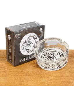 The Bulldog Amsterdam Glas-Aschenbecher mit Verpackung, transparent mit schwarzem Bulldoggen-Logo, auf Holztisch.