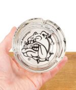 The Bulldog Amsterdam Glas-Aschenbecher in einer Hand gehalten, mit sichtbarem Logo und Zigarrenmulden.