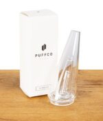 Klarer Puffco Peak Glasaufsatz neben der Originalverpackung auf Holztisch.