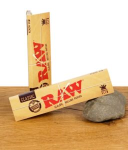 Zwei Packungen RAW Classic Kingsize Slim Rolling Papers, mit Naturfaserband umwickelt, auf einem Stein liegend.