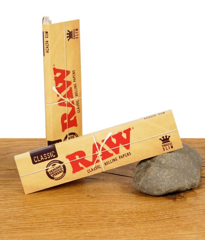 Zwei Packungen RAW Classic Kingsize Slim Rolling Papers, mit Naturfaserband umwickelt, auf einem Stein liegend.