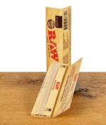Geöffnete RAW Classic Kingsize Slim Packung mit einem einzelnen Rolling Paper, stehend auf einem Stein.