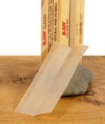 Einzelnes RAW Classic Kingsize Slim Rolling Paper aufgestellt vor der Verpackung, mit Wasserzeichenstruktur sichtbar.