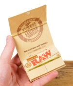 Geöffnete RAW Artesano Organic Hemp Packung mit Papers, Tips und abnehmbarer Mischungsschale in einer Hand gehalten.