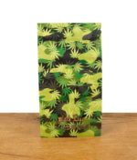 Der Black Leaf Zipperbeutel mit Hanfblatt-Camouflage-Design steht aufrecht auf einer Holzoberfläche. Das Black Leaf-Logo und der Schriftzug „In Weed We Trust“ sind sichtbar.