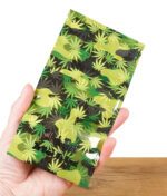 Eine Hand hält einen Black Leaf Zipperbeutel mit grünem Camouflage-Design aus Hanfblättern. Das Black Leaf-Logo und der Schriftzug „In Weed We Trust“ sind aufgedruckt.