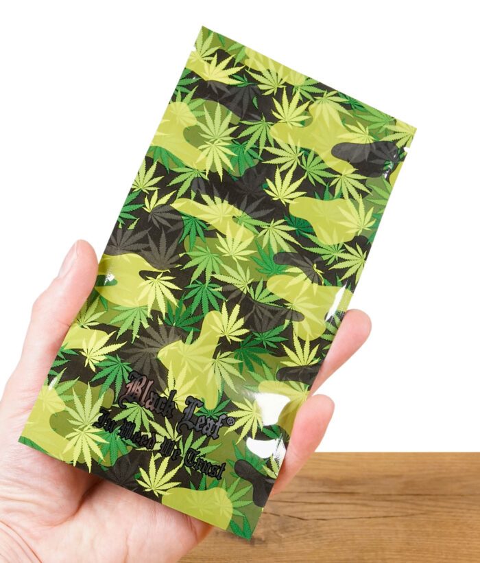 Eine Hand hält einen Black Leaf Zipperbeutel mit grünem Camouflage-Design aus Hanfblättern. Das Black Leaf-Logo und der Schriftzug „In Weed We Trust“ sind aufgedruckt.