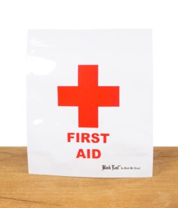 Ein Black Leaf Zipperbeutel mit „First Aid“-Aufdruck und rotem Kreuz steht auf einer Holzoberfläche.
