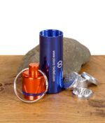 Storz & Bickel Kapselbehälter in Blau mit orangem Deckel, Schlüsselring und Dosierkapseln auf Holz mit Stein im Hintergrund.