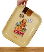 Hand hält das RAW Rolling Tray mit Pin-up-Girl-Design und RAW-Logo, geneigt über einem Holztisch.