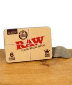 RAW Classic Cone Holder – Metalldose für King Size Cones mit klassischem RAW-Design, platziert auf einem Holztisch mit Stein.