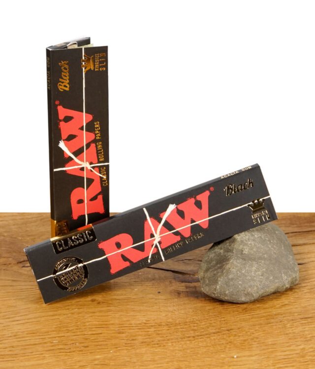 RAW Black Classic Rolling Papers – zwei Packungen ultradünnes, ungebleichtes Zigarettenpapier im schwarzen Design auf Holztisch mit Stein.