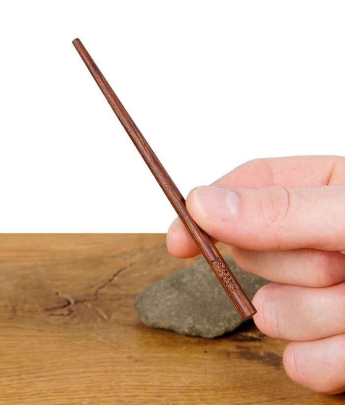 RAW Wooden Poker – dunkelbrauner Holztamper mit RAW-Logo, gehalten zwischen Daumen und Finger vor Holztisch-Hintergrund.