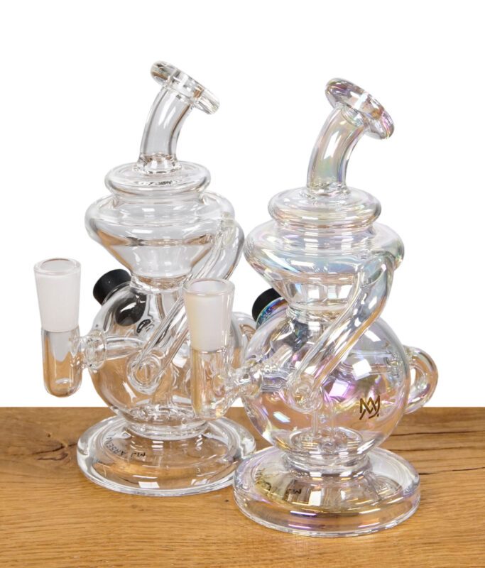 Zwei MJ Arsenal Mini Jig Dab Rigs aus Glas, eine transparente und eine schillernde Variante, nebeneinander auf einem Holztisch platziert.