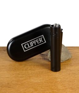 Schwarzes Clipper Metal Feuerzeug stehend vor einer passenden schwarzen Clipper-Metallbox auf einem Holztisch mit Stein im Hintergrund.