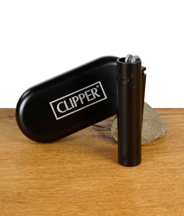 Schwarzes Clipper Metal Feuerzeug stehend vor einer passenden schwarzen Clipper-Metallbox auf einem Holztisch mit Stein im Hintergrund.