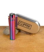 Clipper Feuerzeug in Regenbogenfarben mit silberner Metallbox auf Holztisch, Stein im Hintergrund.