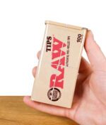 RAW Tin Metalldose in einer Hand gehalten über Holzboden, vor weißem Hintergrund.