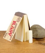 Geöffnetes Heftchen der RAW Perforated Wide Tips mit einzelnem, aufgerolltem Filtertip neben einem Stein auf Holzfläche.