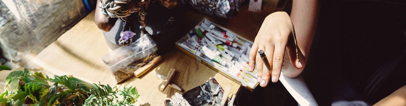 Person hält brennenden Cannabis-Joint in der Hand, daneben Kunstutensilien, Skulpturen und Pflanzen – Symbolbild für die Verbindung von Cannabis und Kunst.