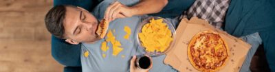 Mann liegt entspannt auf dem Sofa, isst Pizza und Nachos mit einem Getränk in der Hand – typische Szene für Heißhunger nach dem Kiffen.