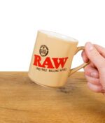 RAW Tasse in beige mit rotem Logo wird in der Hand gehalten
