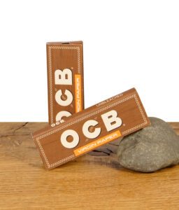 Zwei geschlossene Packungen OCB Virgin Paper Regular Size ungebleicht auf Holztisch mit Stein zur Stabilisierung