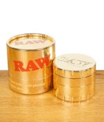RAW Ethereal Grinder goldfarben neben glänzender Originalverpackung – beides stehend auf Holztisch