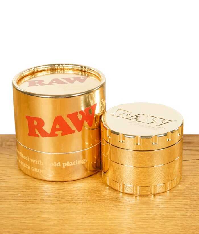 RAW Ethereal Grinder goldfarben neben glänzender Originalverpackung – beides stehend auf Holztisch