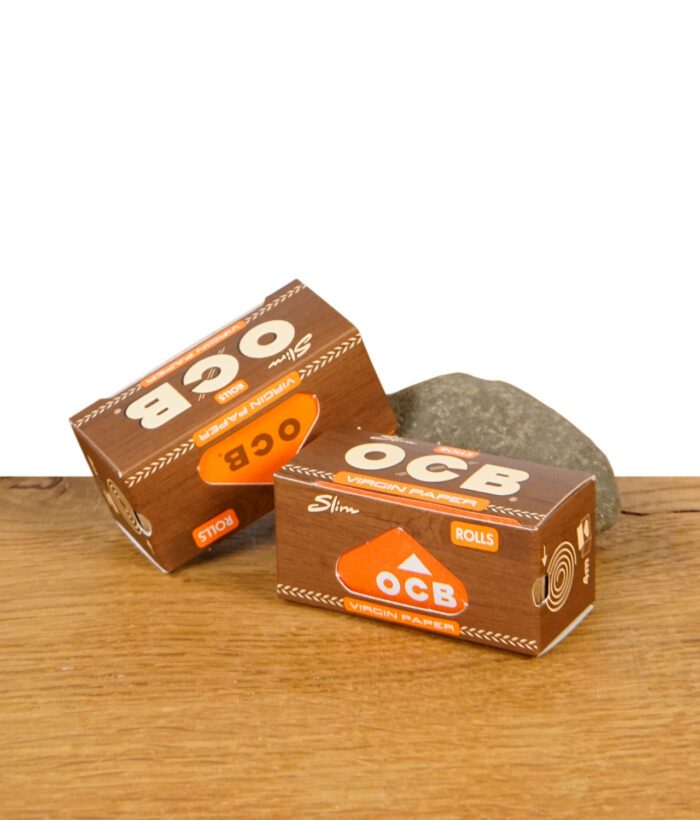 Zwei geschlossene Packungen OCB Virgin Paper Rolls Slim auf einem Holzuntergrund mit Stein