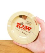 Hand hält den runden RAW Magnet-Aschenbecher mit beige-rotem Design und Kordelaufdruck