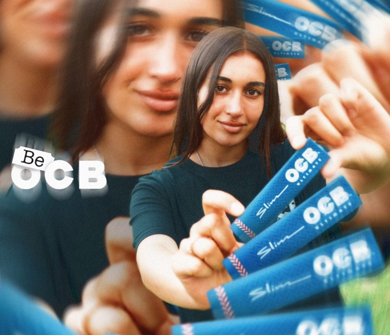 Junge Frau hält mehrere OCB Ultimate Slim Papers in die Kamera, daneben ist der Schriftzug „Be OCB“ zu sehen
