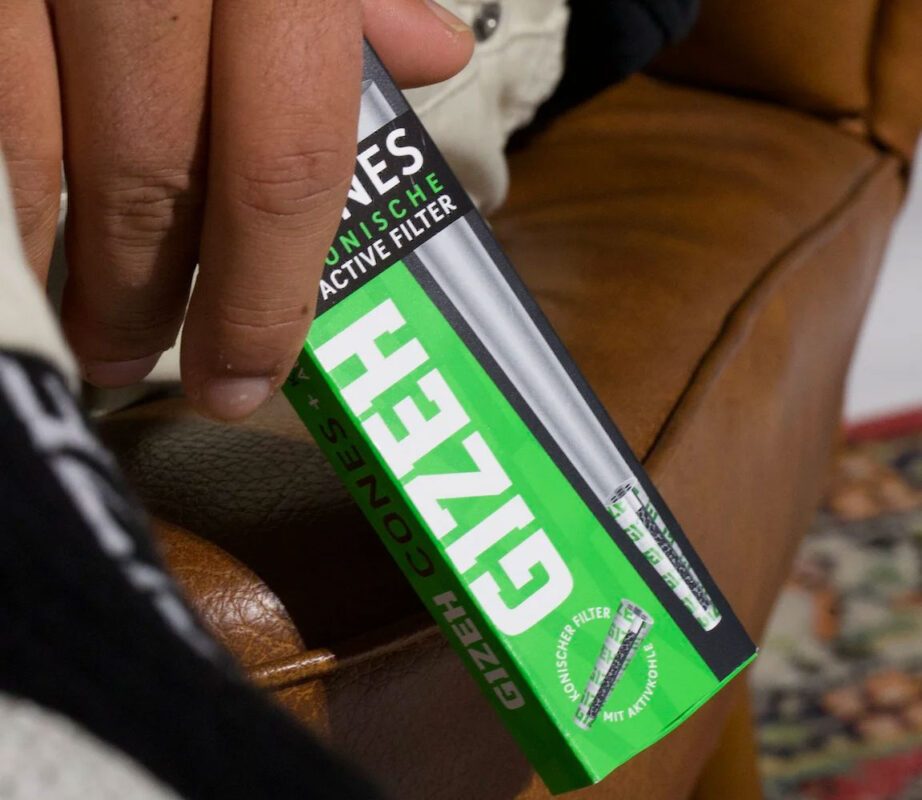 Person hält eine grüne Packung GIZEH Cones mit konischem Aktivkohlefilter in der Hand – Nahaufnahme auf einem braunen Ledersessel.