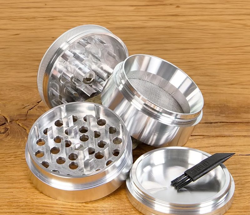 Geöffneter RAW Pollinator Aluminium Grinder mit Sieb und Reinigungsbürste auf Holzoberfläche