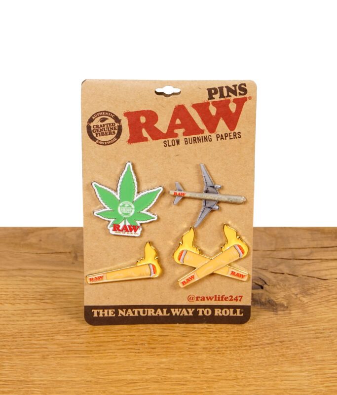 RAW Pin Set V2 auf brauner Verpackungskarte mit vier Pins in den Motiven Hanfblatt, Joint-Flammen, gekreuzte Joints und RAW-Flugzeug.