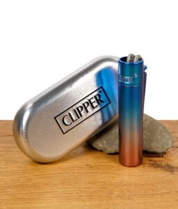 Clipper Metallfeuerzeug im Dawn Gradient Look in Blau-Rot-Farbverlauf vor silberner Clipper-Metallbox auf Holzuntergrund