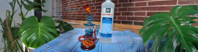 Dabbing Rig aus buntem Glas neben Isopropanol-Reiniger auf Tisch mit Pflanze