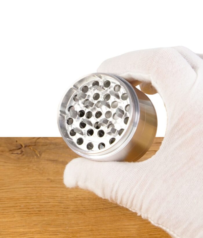 RAW Aluminium Grinder mit Mahlzähnen, in einer Hand mit Handschuh gehalten, Nahaufnahme.